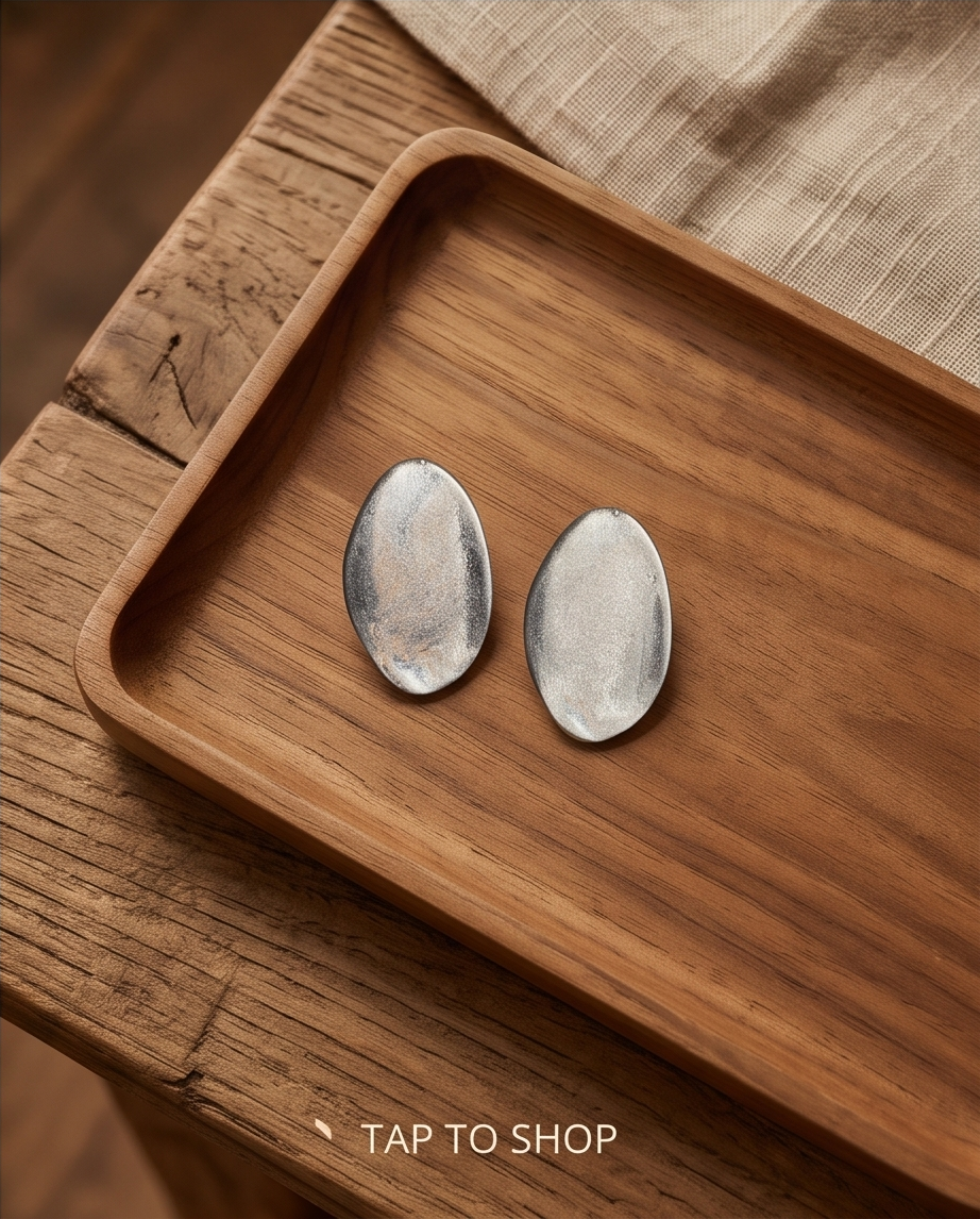 Pair of silver tone brushed oval disc stud earrings standing on warm walnut wood in dappled sunlight