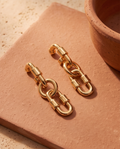 Ear close up of model wearing gold tone chunky interlocking oval chain link drop earring with potted plant and warm Mediterranean courtyard background