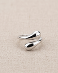 Model's hand resting on wooden chair arm wearing 925 silver plated sculptural double teardrop wrap open adjustable ring with cream satin sleeve