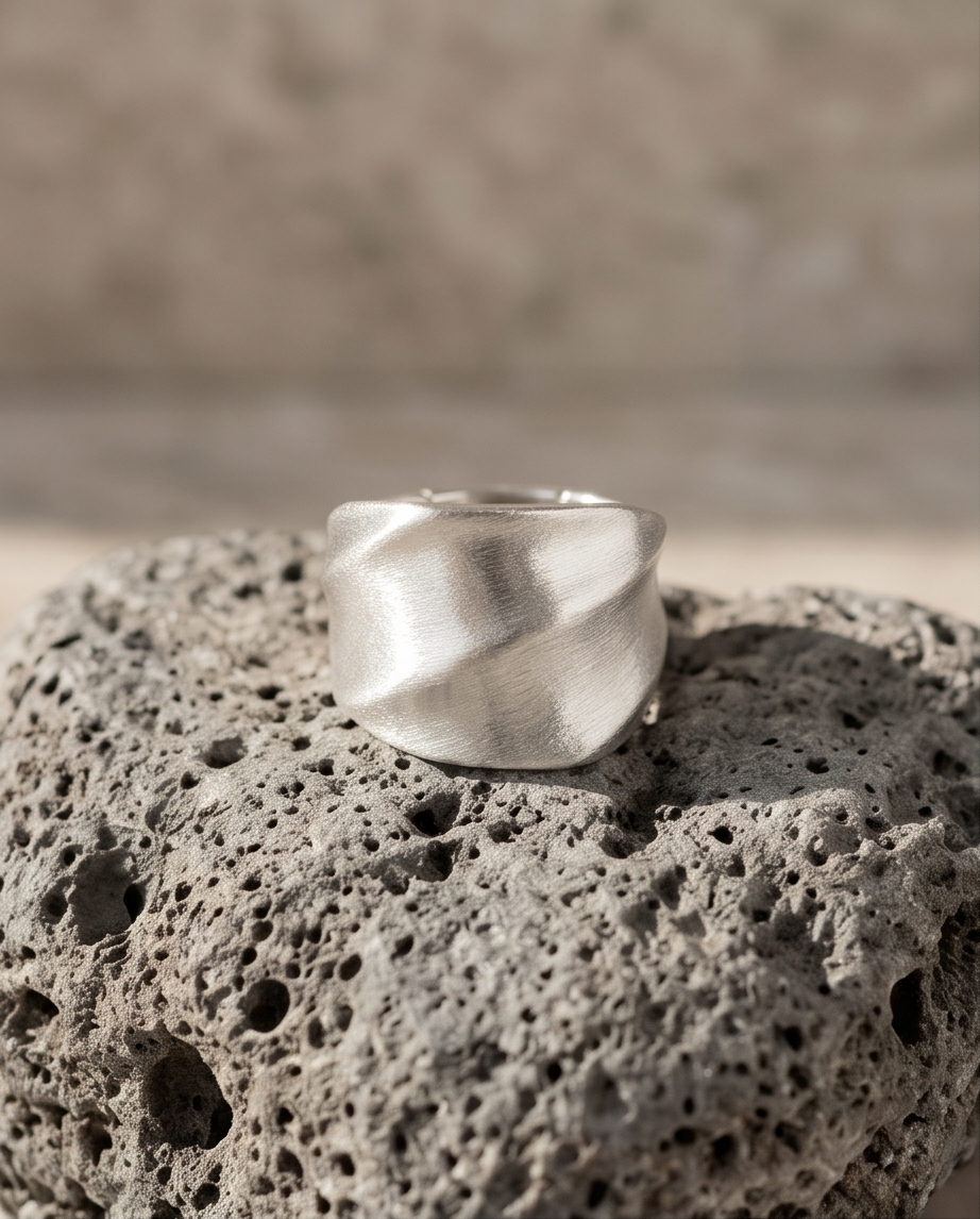 925 silver plated brushed dome sculptural twisted facet wide band ring balanced on volcanic pumice rock in warm sandy light showing full dome profile and diagonal twist facet planes