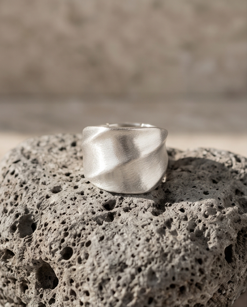 925 silver plated brushed dome sculptural twisted facet wide band ring balanced on volcanic pumice rock in warm sandy light showing full dome profile and diagonal twist facet planes