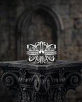 Silver plated gothic fleur de lis baroque cross CZ pave band open adjustable ring standing on ornate dark carved stone column capital in front of gothic cathedral arch — dark luxury editorial product shot