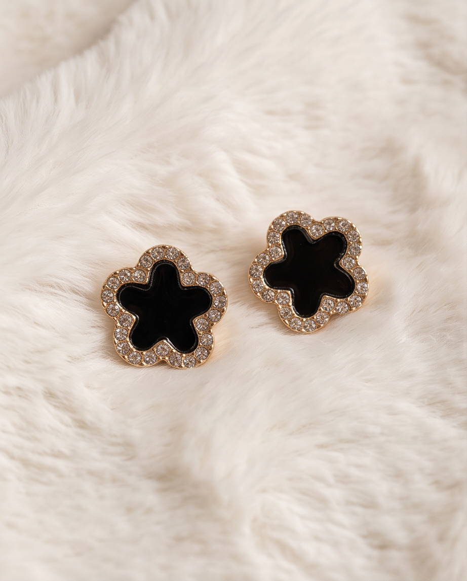 Pair of gold tone black enamel clover CZ pave stud earrings laid on cream white fluffy fur fabric showing full clover silhouette with sparkling cubic zirconia pave border and deep black enamel centre