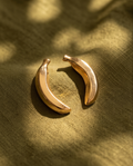 Gold sculptural crescent stud earrings flat lay on olive linen fabric in dappled sunlight