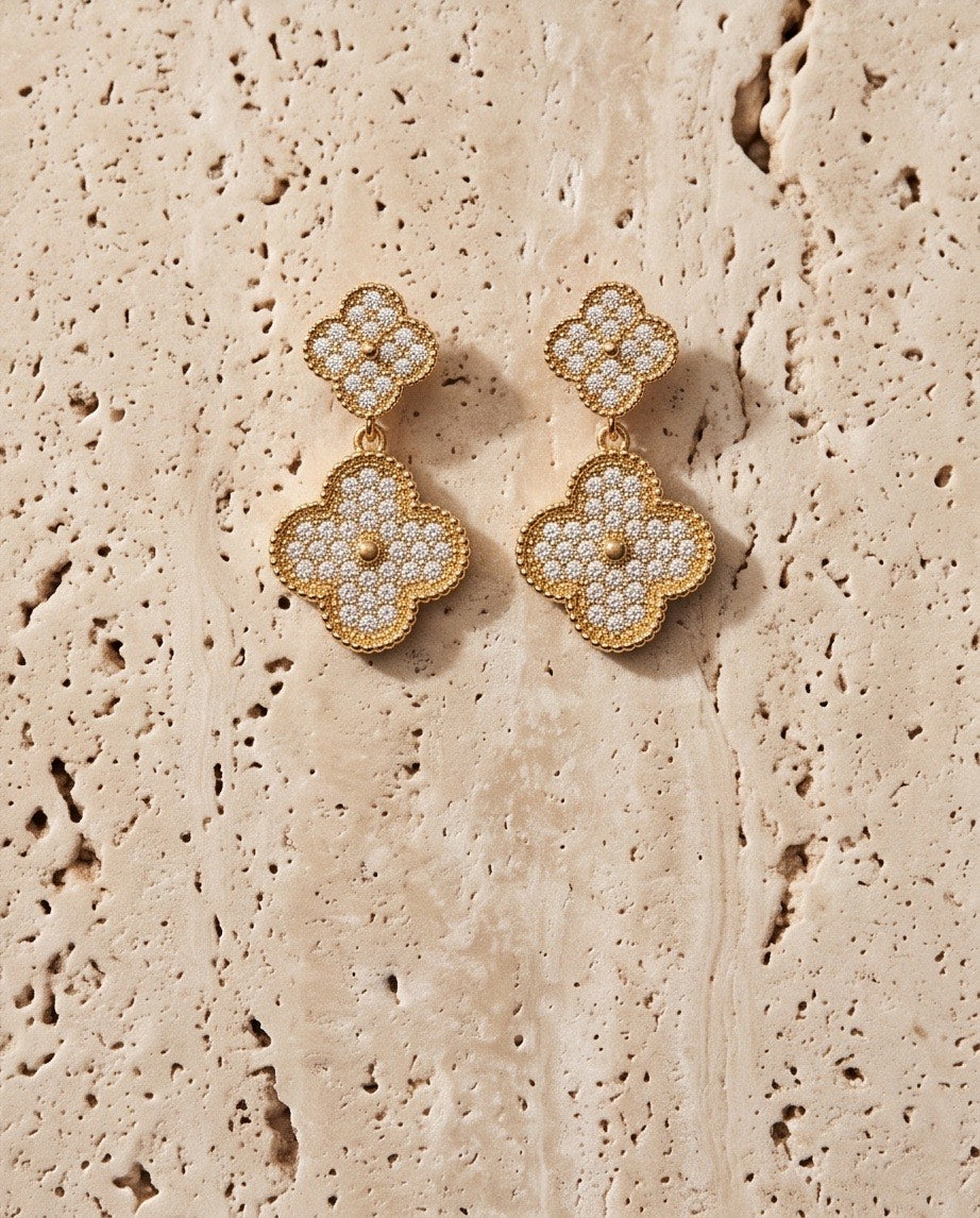 Gold pavé crystal double clover drop earrings flat lay on travertine stone surface