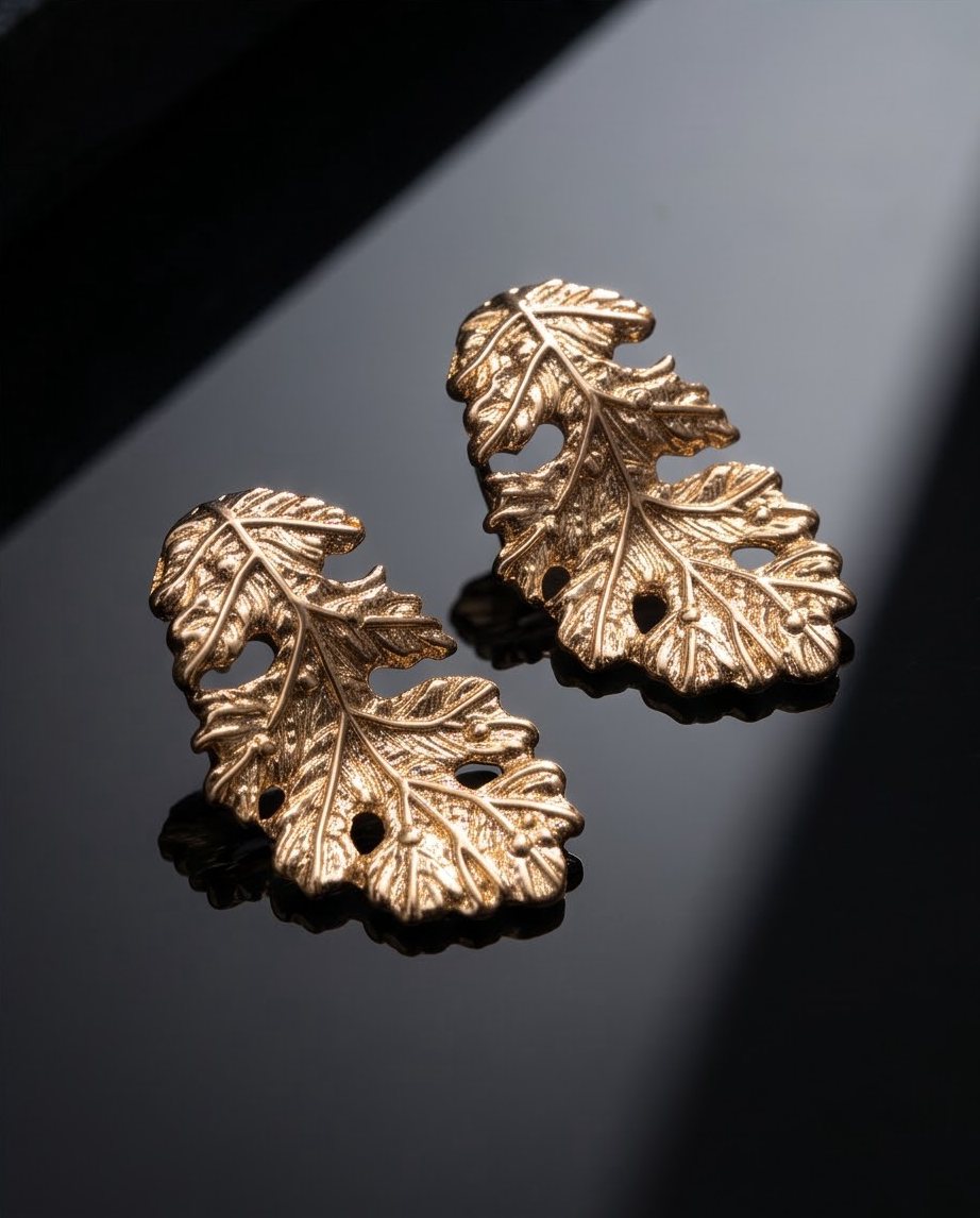 Gold botanical leaf stud earrings pair on dark reflective surface with light and shadow