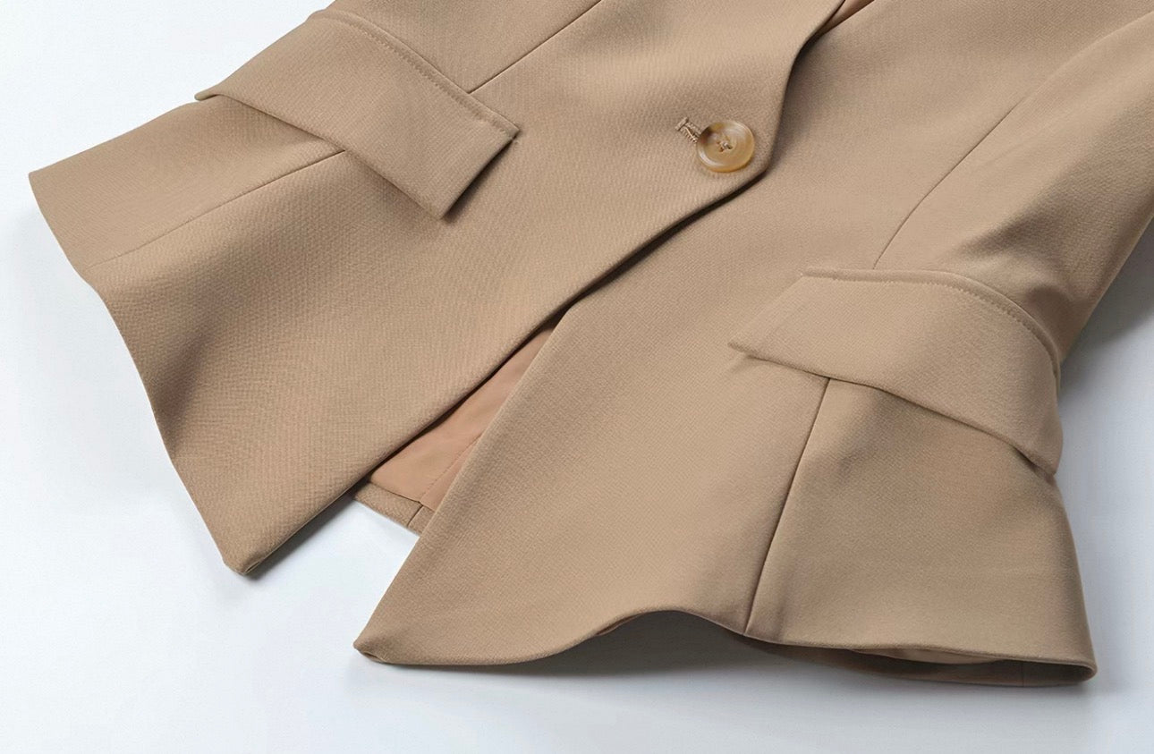 A close-up of a beige tailored jacket or blazer with structured lapels, a single visible button, and a textured fabric, photographed on a plain light background.