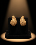 Pair of gold glitter teardrop stud earrings on champagne silk fabric next to wax-sealed envelope and opera glasses
