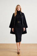 A woman models a black knit turtleneck dress with a matching black cape jacket featuring gold buttons, a black belt, and brown pointed-toe heels, set against a neutral studio background.