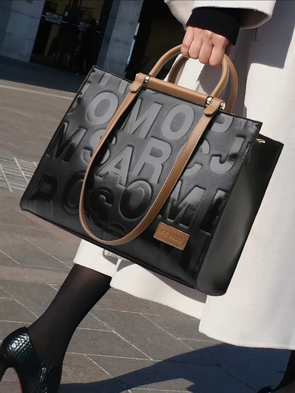 A black tote bag with embossed lettering and tan leather handles, carried by a person wearing a white coat and black tights in an outdoor urban setting.