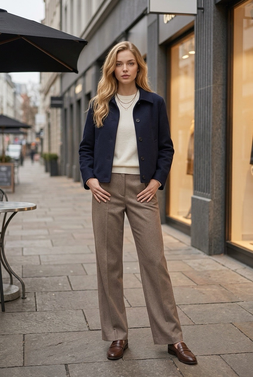 A woman stands on a city sidewalk wearing a navy blue jacket, cream sweater, taupe wide-leg trousers, and brown loafers, with storefronts and a street scene in the background.