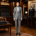 A woman stands in a luxury tailoring room wearing a light gray two-piece suit with a white shirt and dark gray turtleneck. The background features dark wood cabinets, fabric rolls, and a dress form displaying a navy suit.
