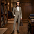 A woman stands in a warm, wood-paneled boutique wearing a tailored light gray suit with a white turtleneck, holding a dark brown handbag. A sign on the wall reads 'WISTERIA & CO.