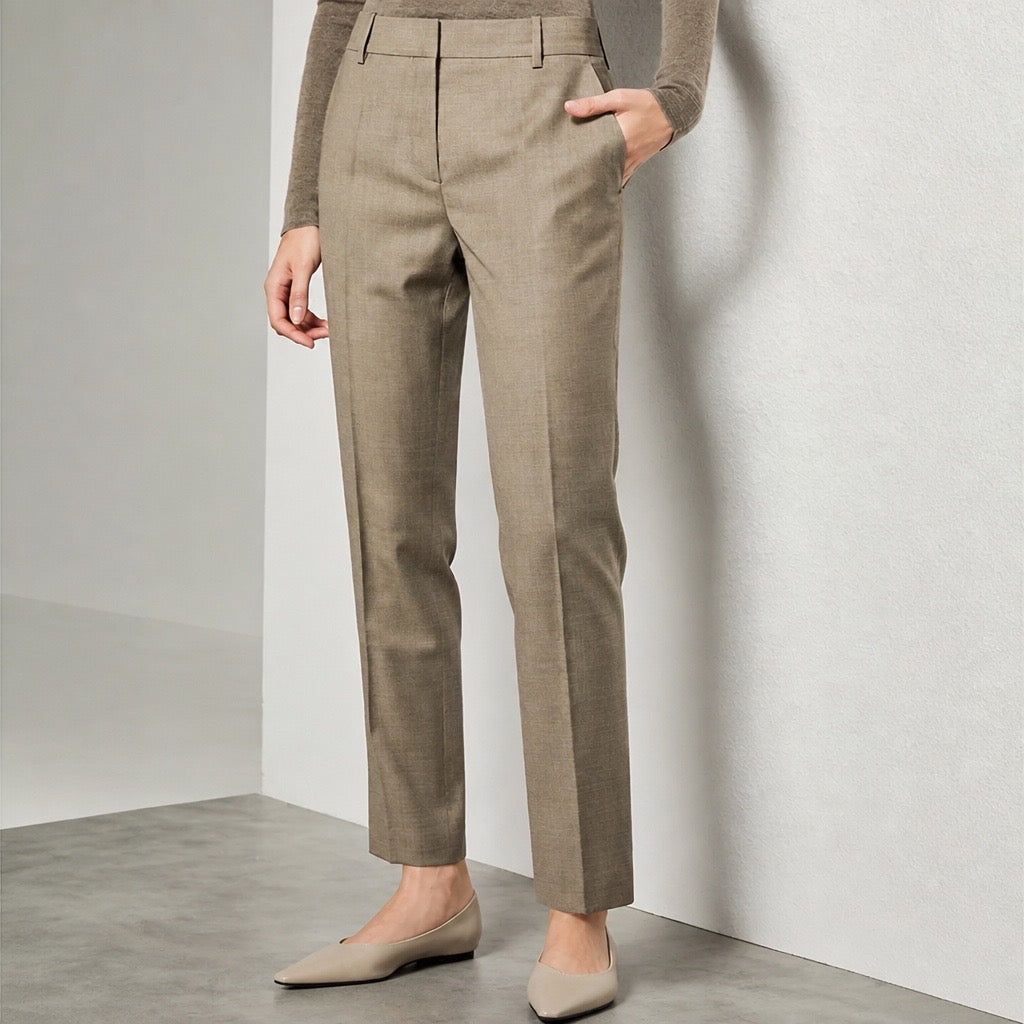 A model wearing high-waisted, straight-leg trousers in a taupe-beige color, paired with a matching knit top and beige pointed flats, standing against a minimalist light gray wall.