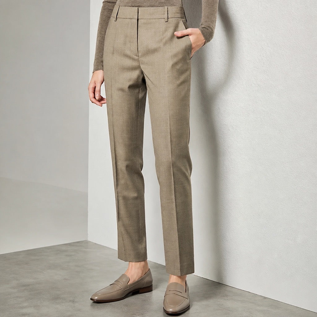 A person wearing tailored straight-leg trousers in a warm taupe-beige color, paired with a matching long-sleeve top and loafers, standing against a light gray studio wall.
