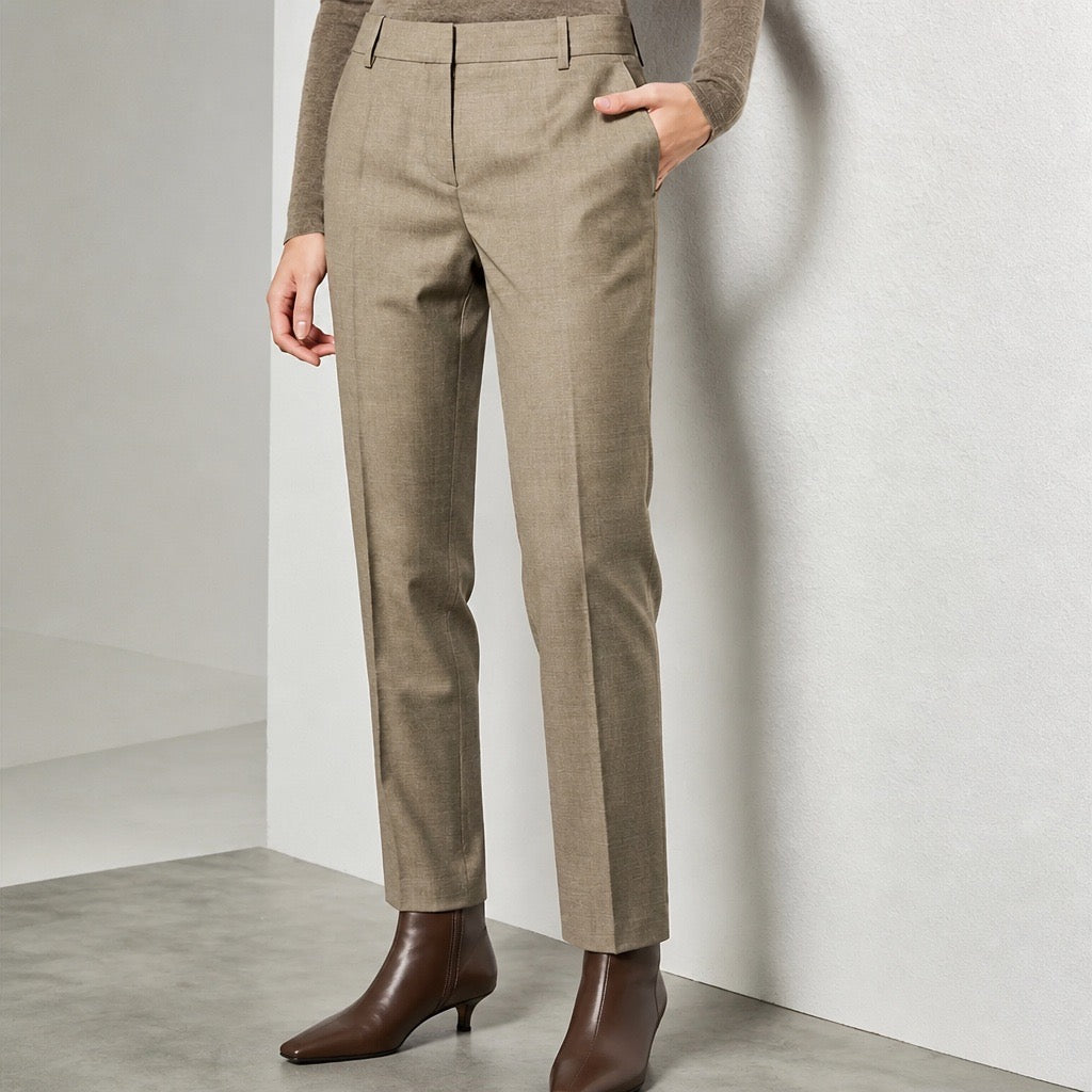 A model wearing straight-leg taupe trousers, a brown knit top, and brown pointed-toe ankle boots, standing against a minimalist light gray wall.