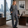 A woman stands in a modern office wearing a tailored gray pinstripe blazer and matching wide-leg trousers over a gray top. Large windows behind her show a city skyline, and a desk with computers and framed wall art are visible.