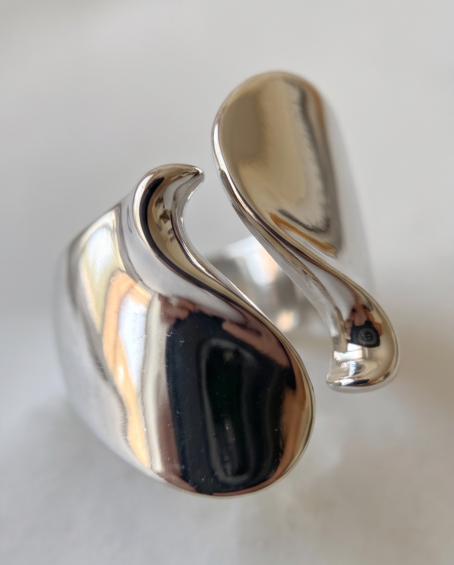 Macro close up of silver tone high polish sculptural S-wave open adjustable ring on white surface showing liquid mercury mirror finish and wave detail