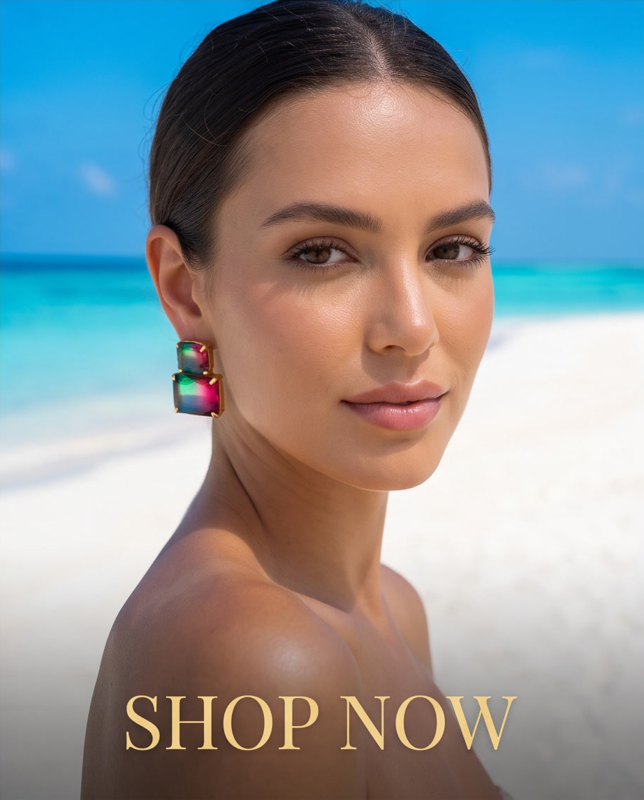 Gold-tone watermelon crystal double drop stud earrings laid on white textured sand with detail of faceted gems