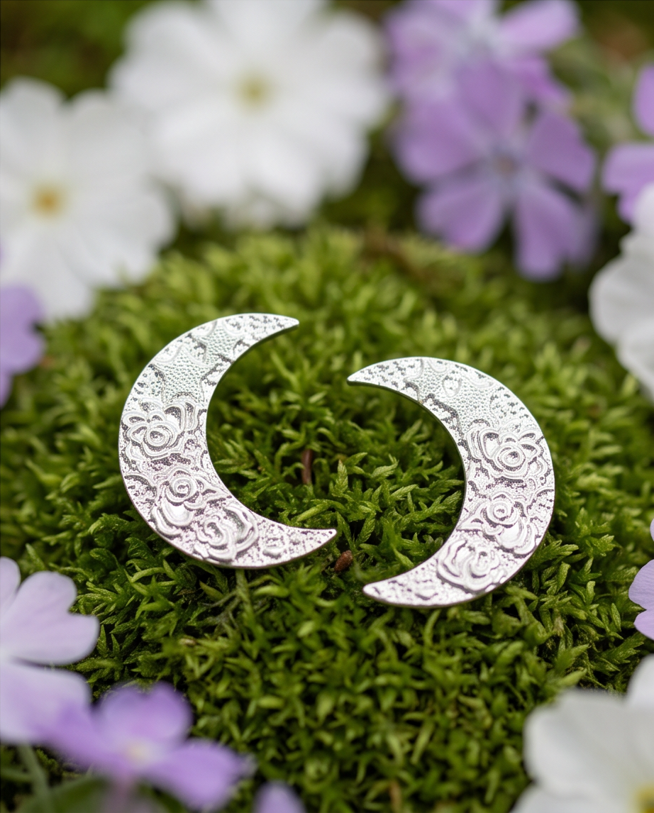 Pair of silver tone embossed floral crescent moon stud earrings on green moss surrounded by white and purple flowers
